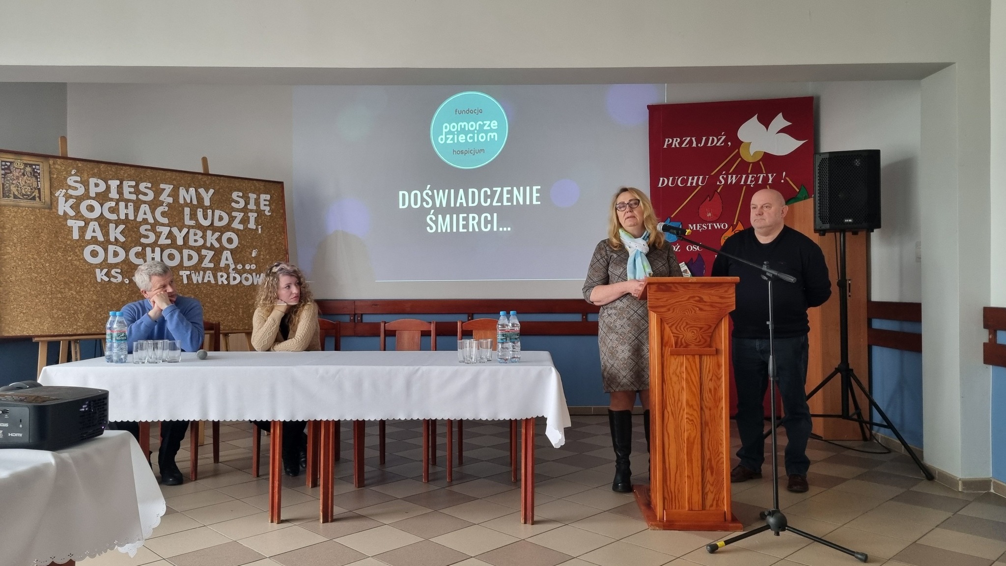 Na stole znajdują się plakaty z hasłami w języku polskim. W tle grupa osób. Na pierwszym planie stoi projektor.