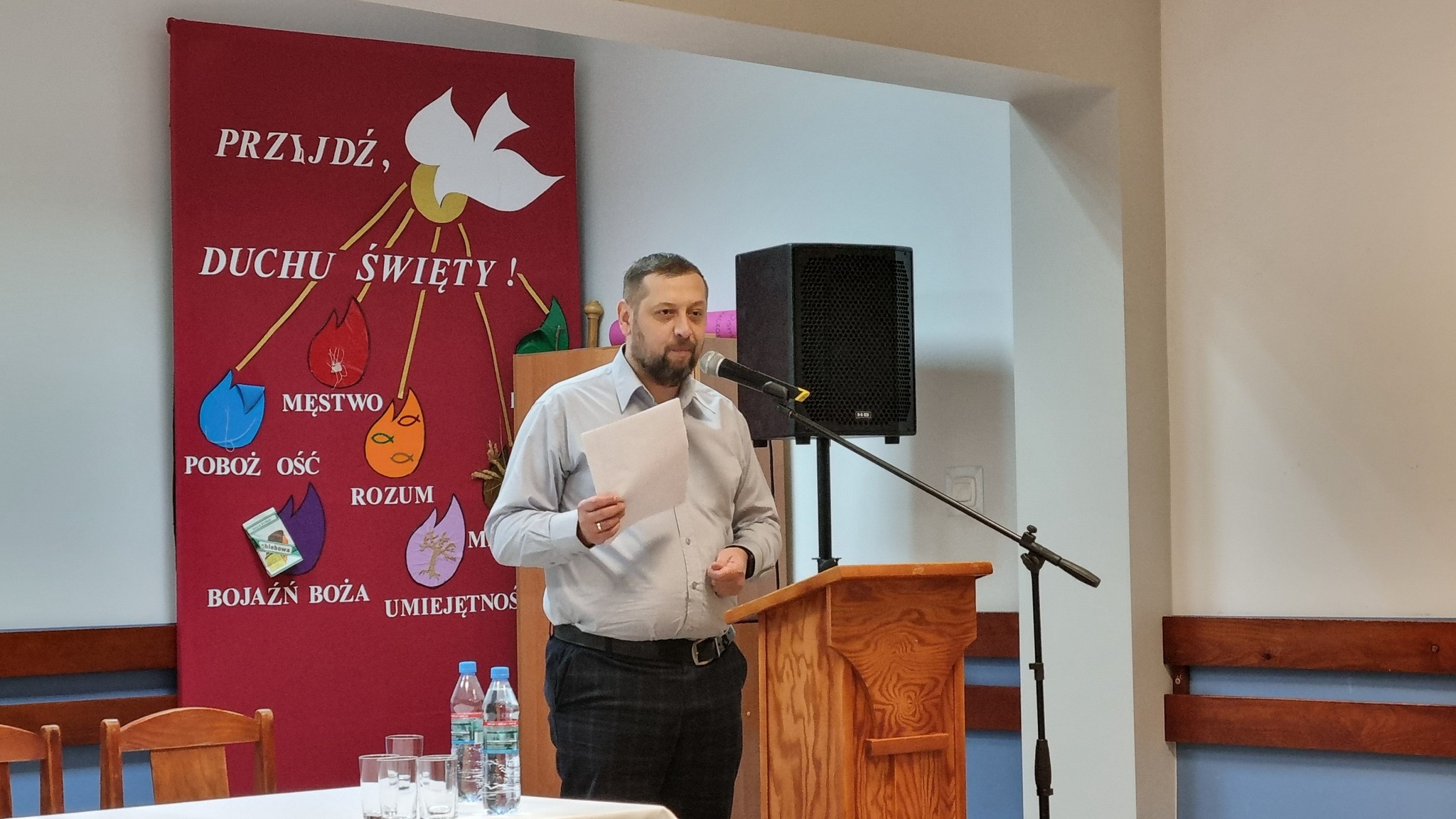 Mężczyzna przemawia podczas wydarzenia. Na plakatach widnieją napisy w języku polskim: „Przyjdź, Duchu Święty!”, „Pobożność Ostatniej Wieczerzy”, „Rozum”, „Boża bojażnia” i „Umiejetność”.