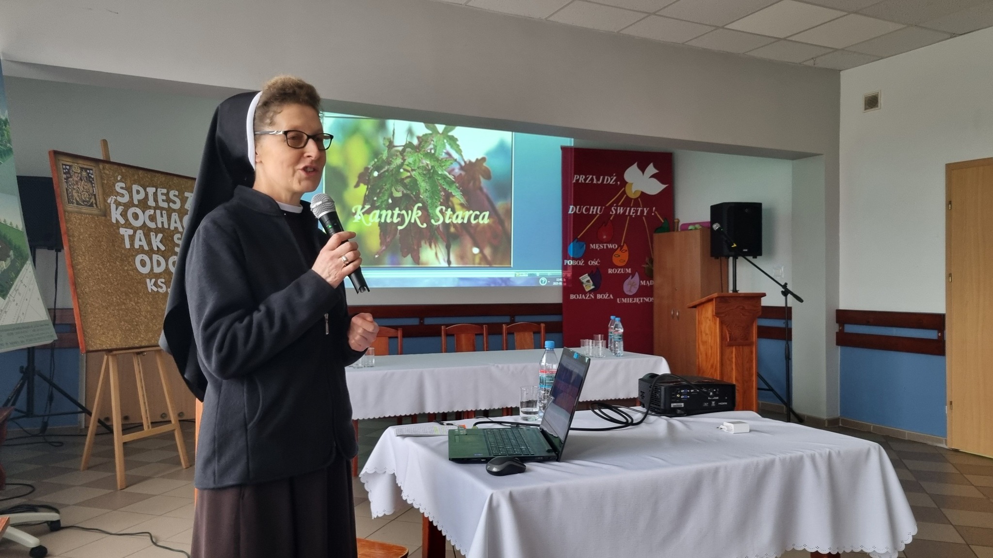 Na stole znajduje się prezentacja multimedialna z wyświetlonym obrazem i tekstem "Kantyk Starca". W tle znajdują się plakaty informacyjne dotyczące wydarzenia. Osoba w habitcie prowadzi prezentację.