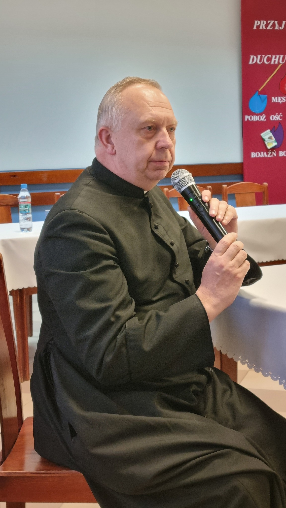 Człowiek w ciemnym, zakrytym płaszczu siedzi przy stole i trzyma mikrofon. W tle widoczny stół z drewnianymi krzesłami oraz plakaty.