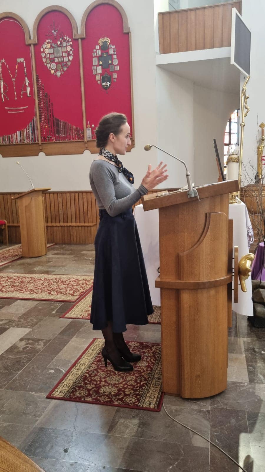 Na zdjęciu widoczna jest osoba stojąca przy drewnianej, kwadratowej pelli. W tle widać wnętrze kościelne z podłogą wyłożoną kilimami oraz dekoracyjne panele i elementy wyposażenia. Na ścianie znajduje się mikrofon.