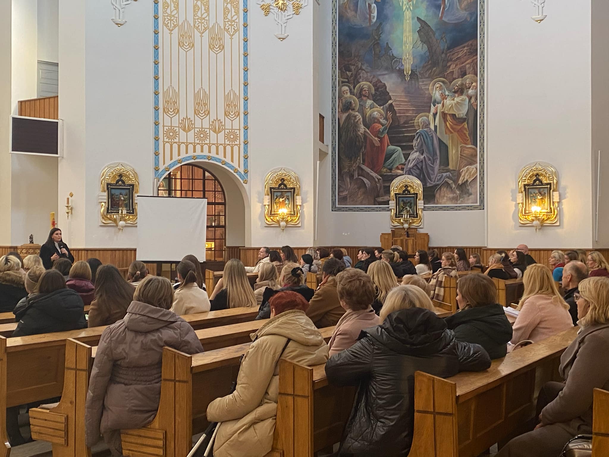 W sali kościelnej zgromadzeni ludzie słuchają prezentacji, na ekranie projekcyjnym wyświetlane są obrazy. W tle znajduje się duży obraz przedstawiający scenę religijną.