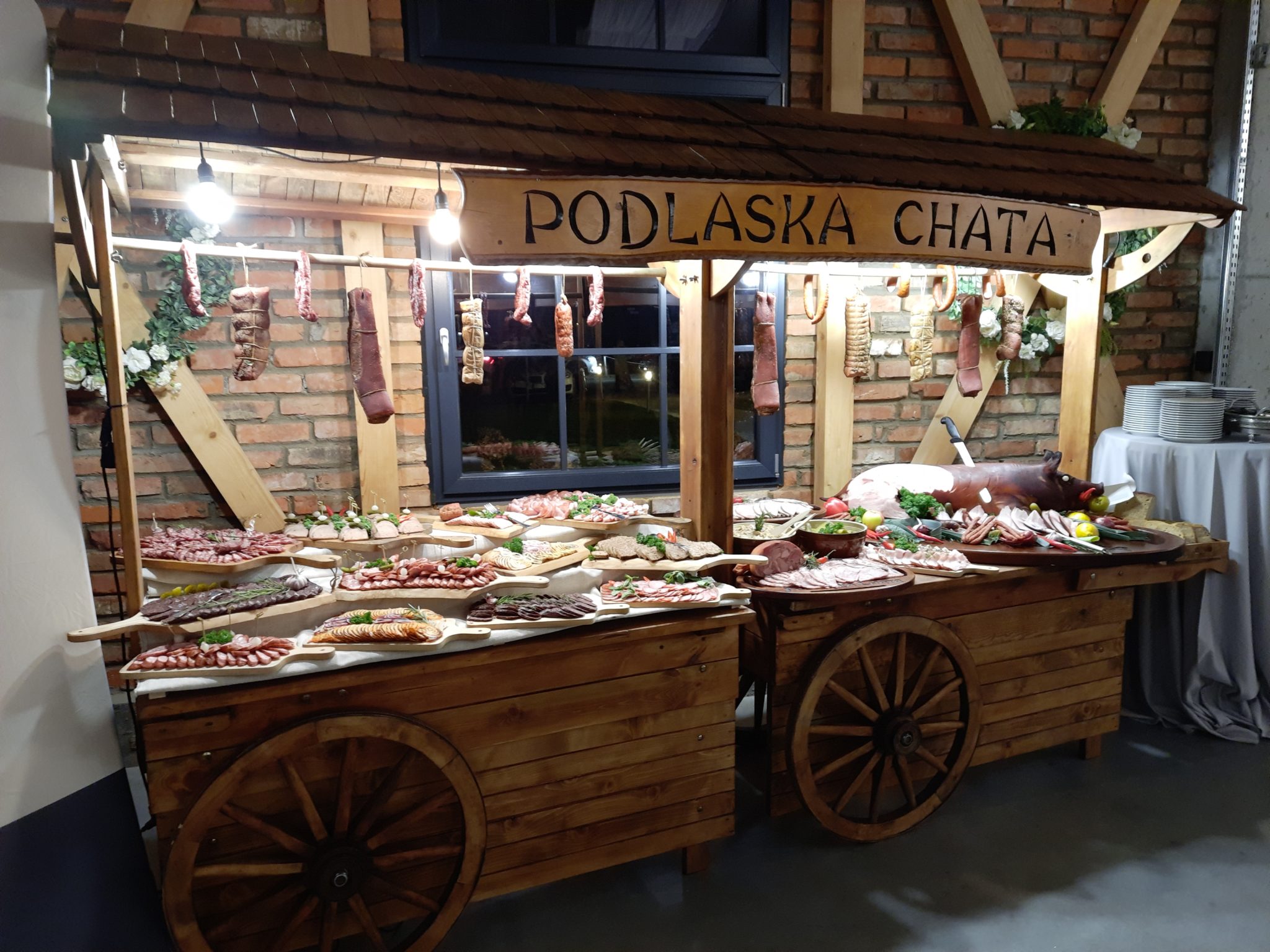 Stół serwujący przekąski i mięsa. Na stole znajdują się drewniane wózki, drewniana konstrukcja z napisem "PODŁASKA CHATA" oraz różne rodzaje potraw, w tym wędliny, sery, owoce i warzywa. W tle ceglane ściany i kwiaty.