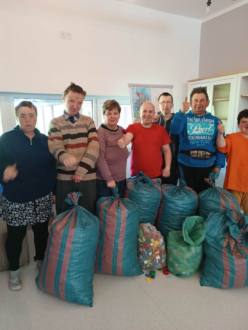 Zdjęcie przedstawia grupę osób zbierających nakrętki plastikowe w workach. W pomieszczeniu znajduje się kilka dużych i małych worków z nakrętkami oraz pojemniki. Zbiórka odbywa się w kwietniu – maju 2023 roku.