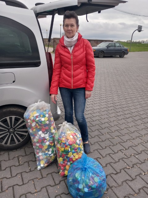 Zdjęcie przedstawia osobę zbierającą nakrętki plastikowe, stojącą obok worków wypełnionych tym materiałem. W tle znajduje się samochód i chodnik z płytkami ceramicznymi.