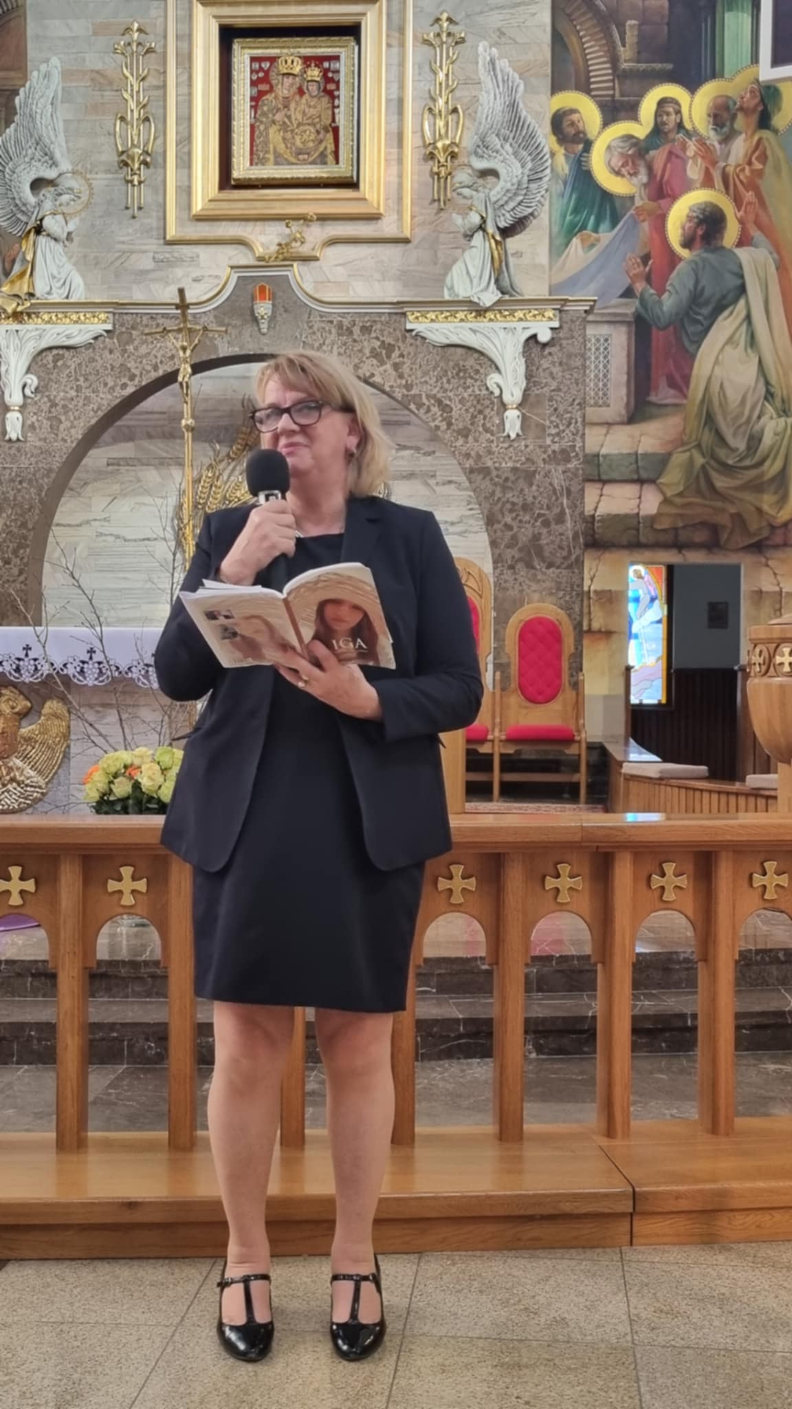 Człowiek w ciemnej marynarce i spódnicy prezentuje przemówienie z mikrofonem wewnątrz kościoła. W tle znajdują się dekoracje świątyni, w tym obrazy i rzeźby. Prezentacja dotyczy postaci biblijnej Judyty.