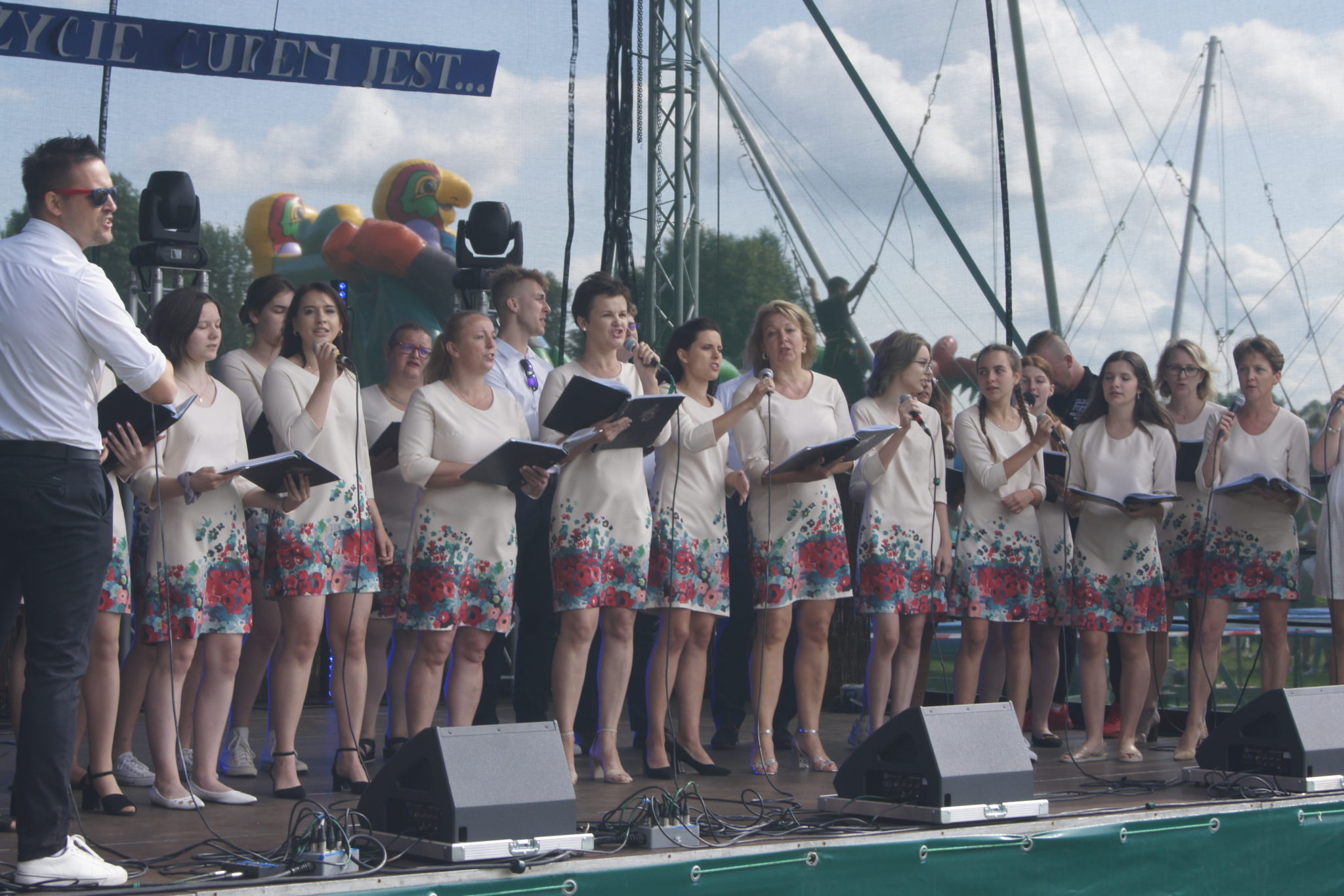 Grupa wokalistów podczas wydarzenia festiwalowego. Na scenie znajduje się mężczyzna i grupa kobiet w kwiatowych spódniczkach, a w tle festiwal z balonami.
