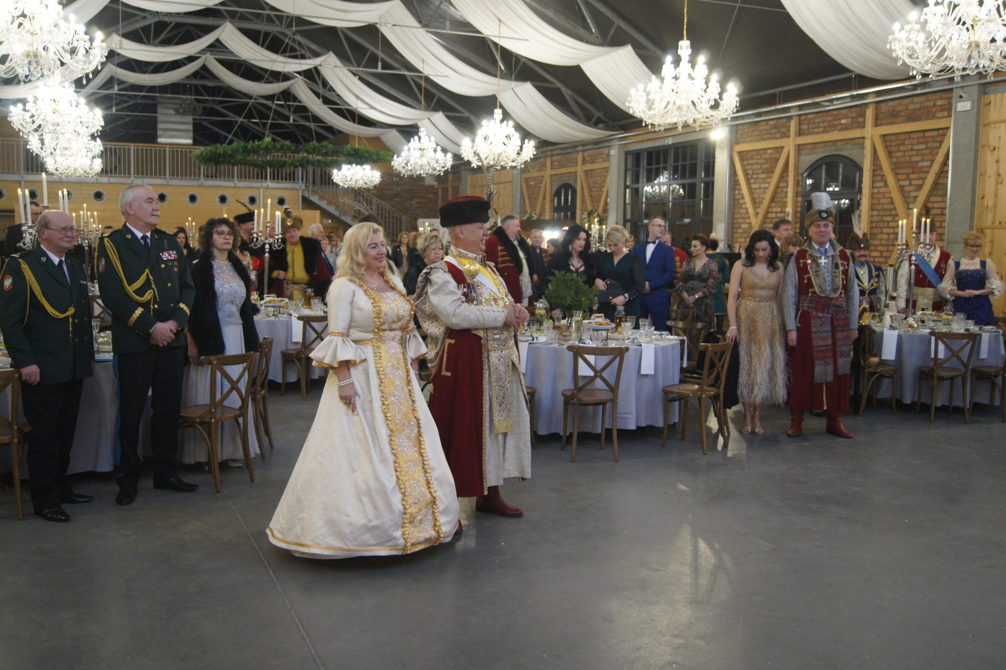 Na zdjęciu widoczna para w tradycyjnych strojach podczas tańca na balu. Otaczają ich inni ludzie ubrani w podobny sposób. W tle znajduje się sala bankietowa z dekoracjami, stołami i oświetleniem.