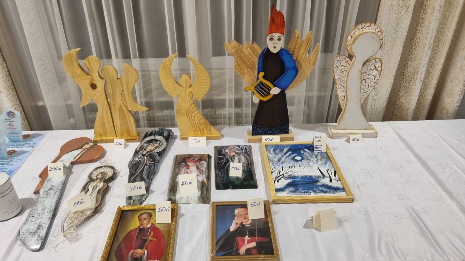 Na stole ustawione są drewniane rzeźby muzyczne i ramki z obrazami, a także tablice z liczbami. W tle widoczny jest element dekoracyjny przypominający łuk.
