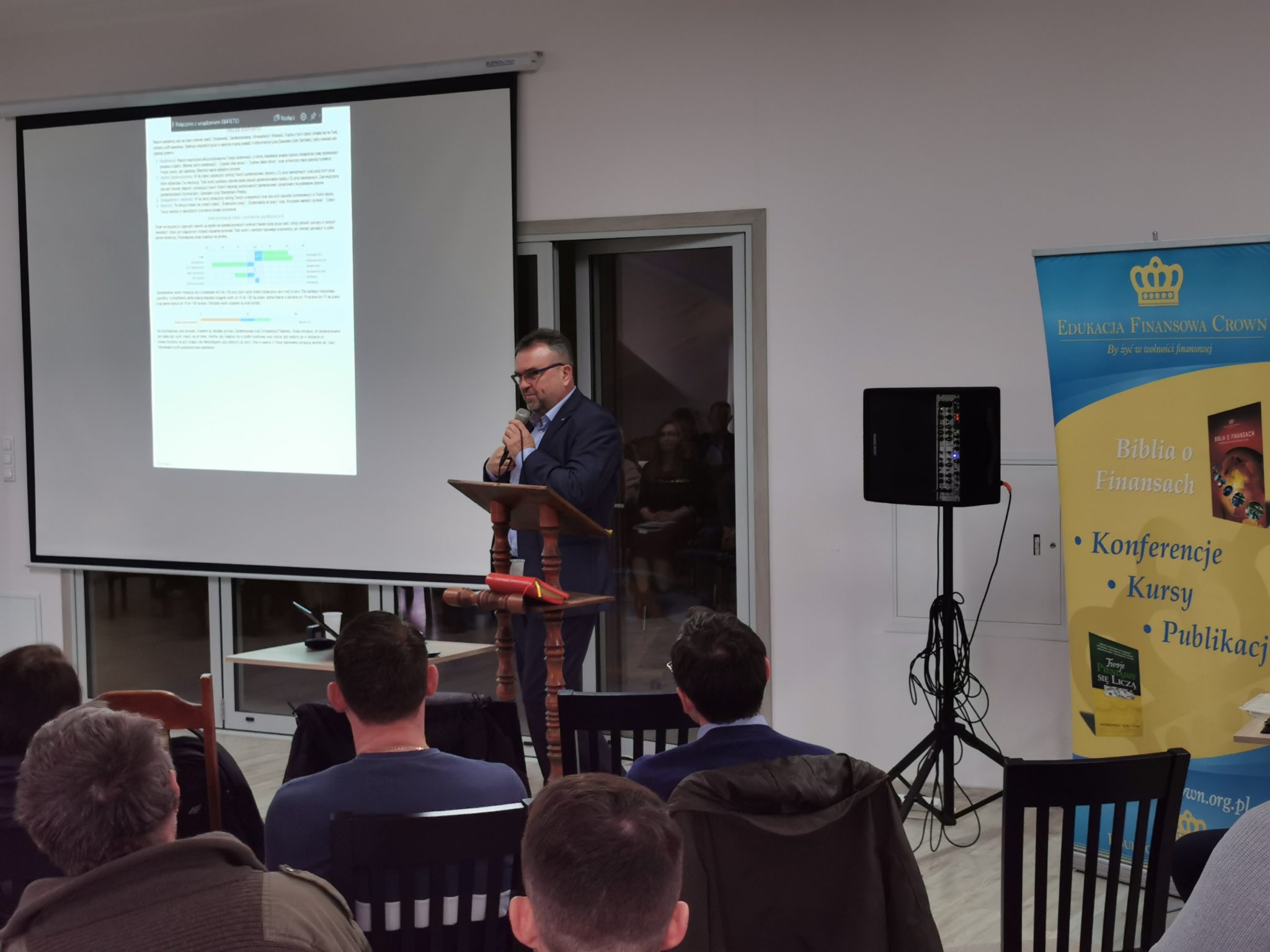 Zdjęcie z wydarzenia. Mężczyzna przemawia do publiczności przed ekranem projekcyjnym, na którym wyświetlany jest tekst. W tle widoczni są słuchacze.