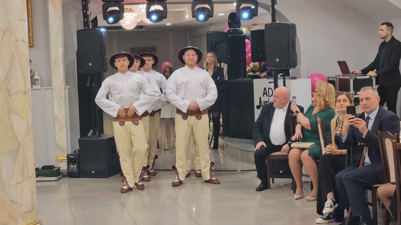 Na scenie grupa mężczyzn w strojach ludowych tańczy, podczas gdy publiczność obserwuje występ. W tle widoczne są głośniki i oświetlenie.