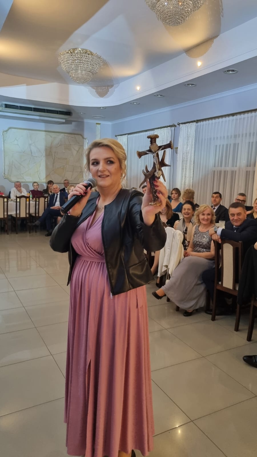 Na zdjęciu widoczna jest osoba w długiej różowej sukni, która trzyma mikrofon i coś, co wygląda jak drewniany krzyż. W tle znajduje się grupa osób siedzących przy stole w sali bankietowej z dekoracjami i lustrami.