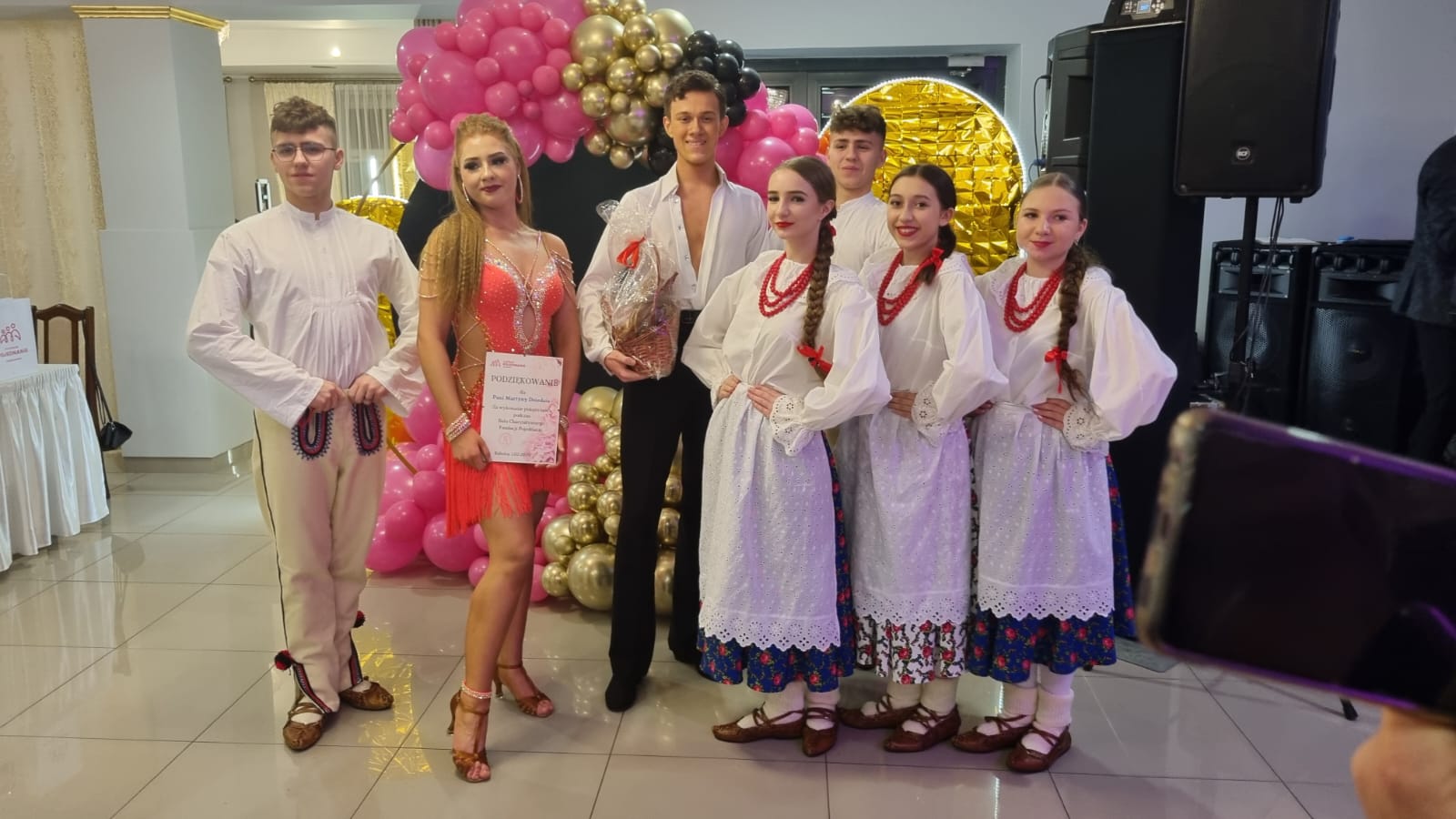 Grupa tancerzy prezentuje się na balu charytatywnym, otoczona dekoracjami balonowymi. Wśród nich widoczny jest mężczyzna w białym garniturze i kobieta w czerwonej sukni. Nagroda leży na podłodze.