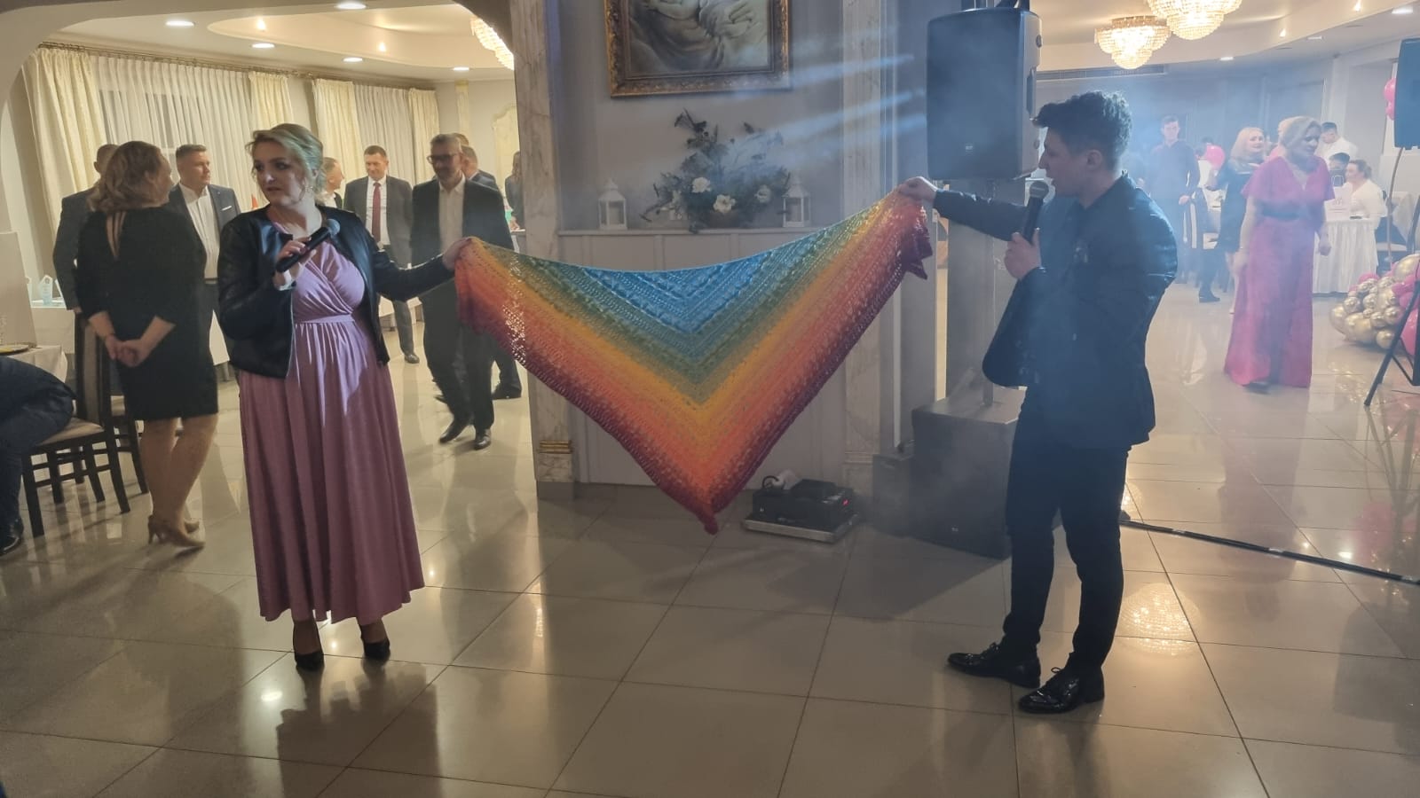 Na zdjęciu widać mężczyznę trzymającego kolorowy szal geometryczny. W tle znajduje się grupa osób ubranych na bal charytatywny, w tym kobieta w różowym makijażu i strój oraz mężczyzna z mikrofonem.