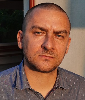 Zdjęcie mężczyzny w szarej koszuli. Mężczyzna patrzy prosto w obiektyw, jego twarz jest wyraźnie oświetlona ciepłym światłem.