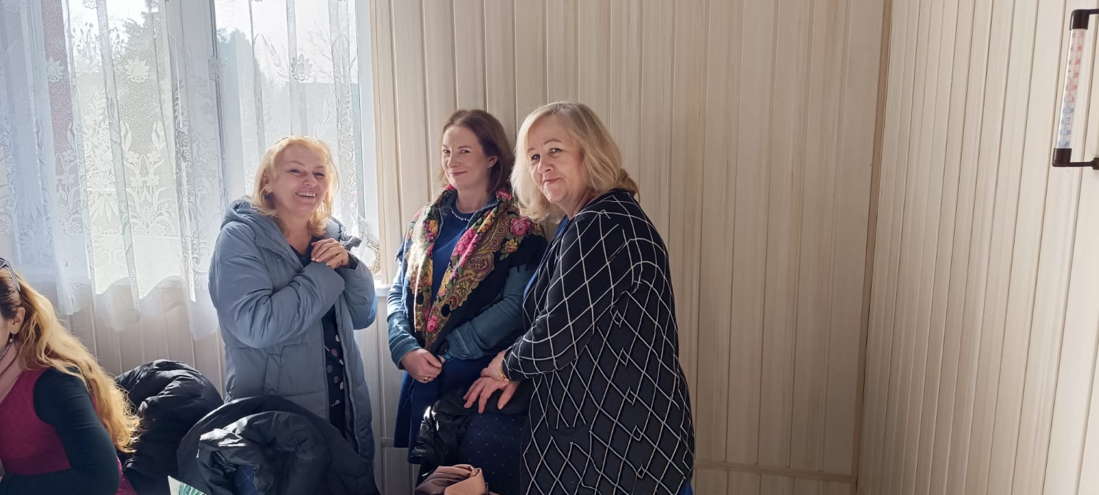 Trzy osoby stoją w pomieszczeniu z oknem. W tle są zasłony i ściana pomalowana na jasnobrązowo. Jedna osoba ma na sobie niebieski płaszcz, druga jest w różowym swetrze, a trzecia w granatowym płaszczyku z geometrycznym wzorem.