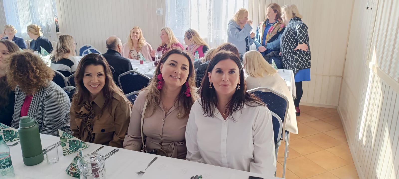 Grupa osób siedzi przy stole w sali konferencyjnej. Trzy kobiety uśmiechają się do kamery. W tle widoczna sala z kilkoma stołami i innymi uczestnikami spotkania. Na stole znajdują się napoje i sztućce.