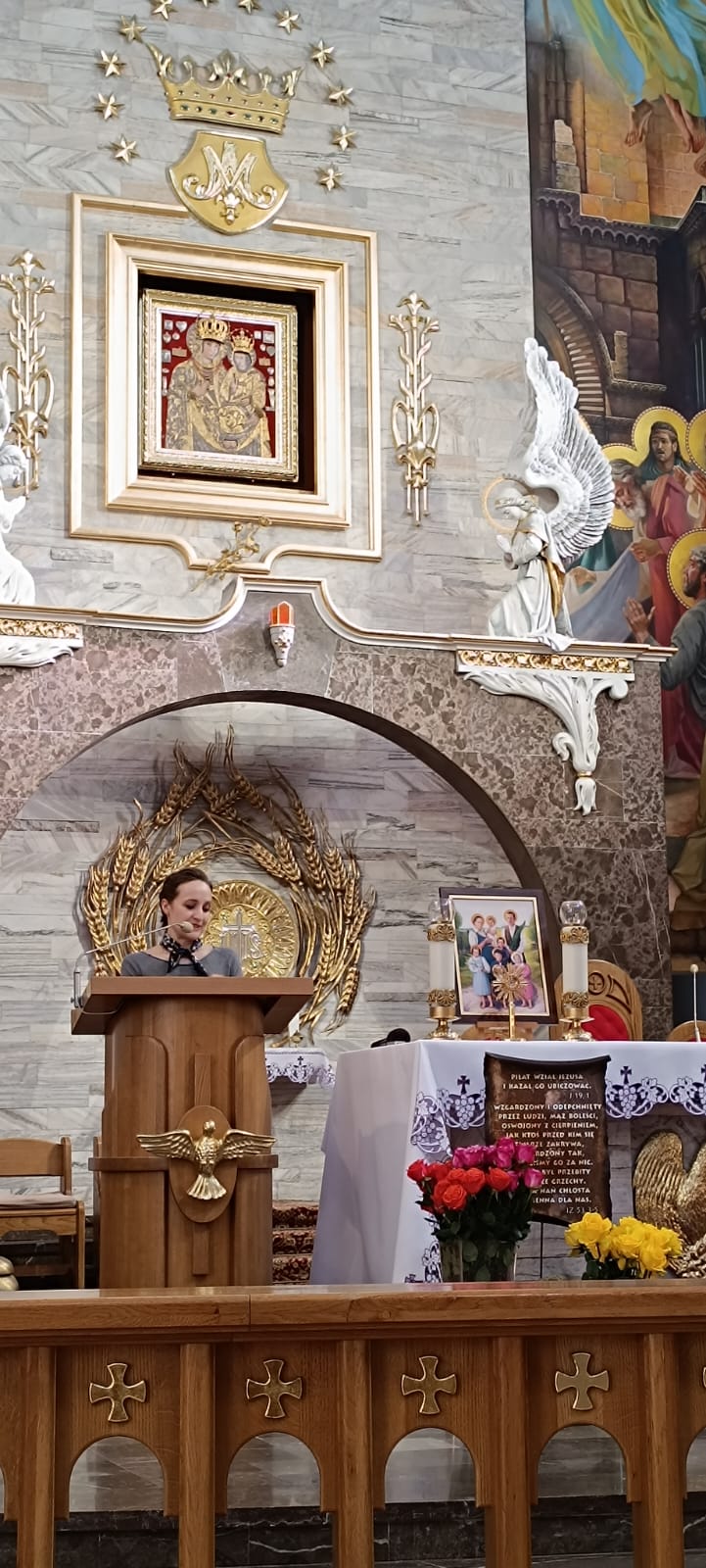 Wnętrze kościoła, na pance stoi osoba z mikrofonem i stołem z wazony. W tle znajduje się obraz Jezusa Chrystusa i anioła oraz ołtarz z obrazem Matki Boskiej.