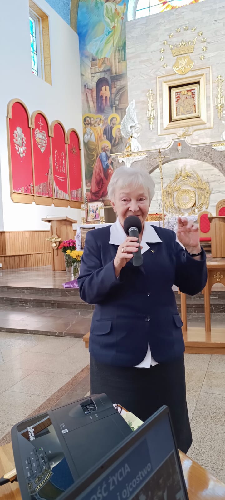 Na zdjęciu widoczna jest osoba w kapeluszniku i szacie, trzymająca mikrofon. W tle znajduje się wnętrze kościelne z ołtarzem i obrazami. Na stole przed osobą stoi urządzenie z wyświetlaczem oraz plakaty.