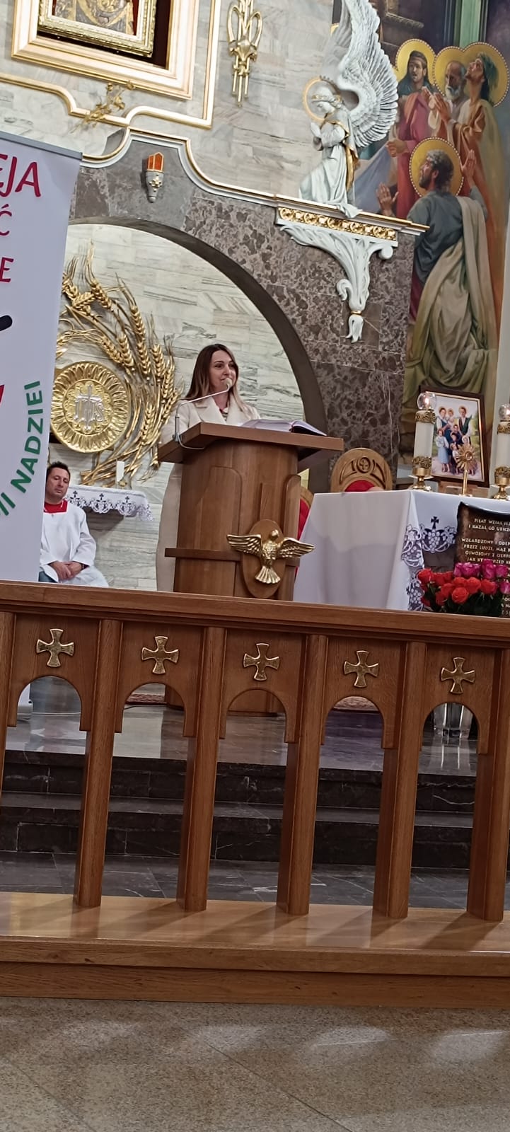 Mężczyzna w białym płaszczu stoi przy scenie z podestem i dekoracjami. W tle znajduje się obraz przedstawiający ptaka oraz napis „NADZIEI”.