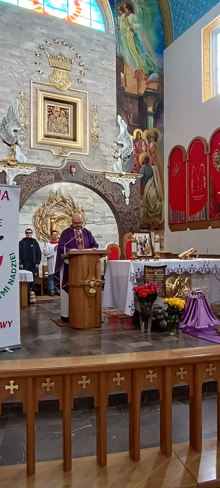 Wnętrze kościoła, na którym stoi osoba w przebraniu kapłana podczas mszy świętej. W tle widoczny ołtarz z dekoracjami i obrazy religijne. Na ławce znajduje się ołtarzewa.