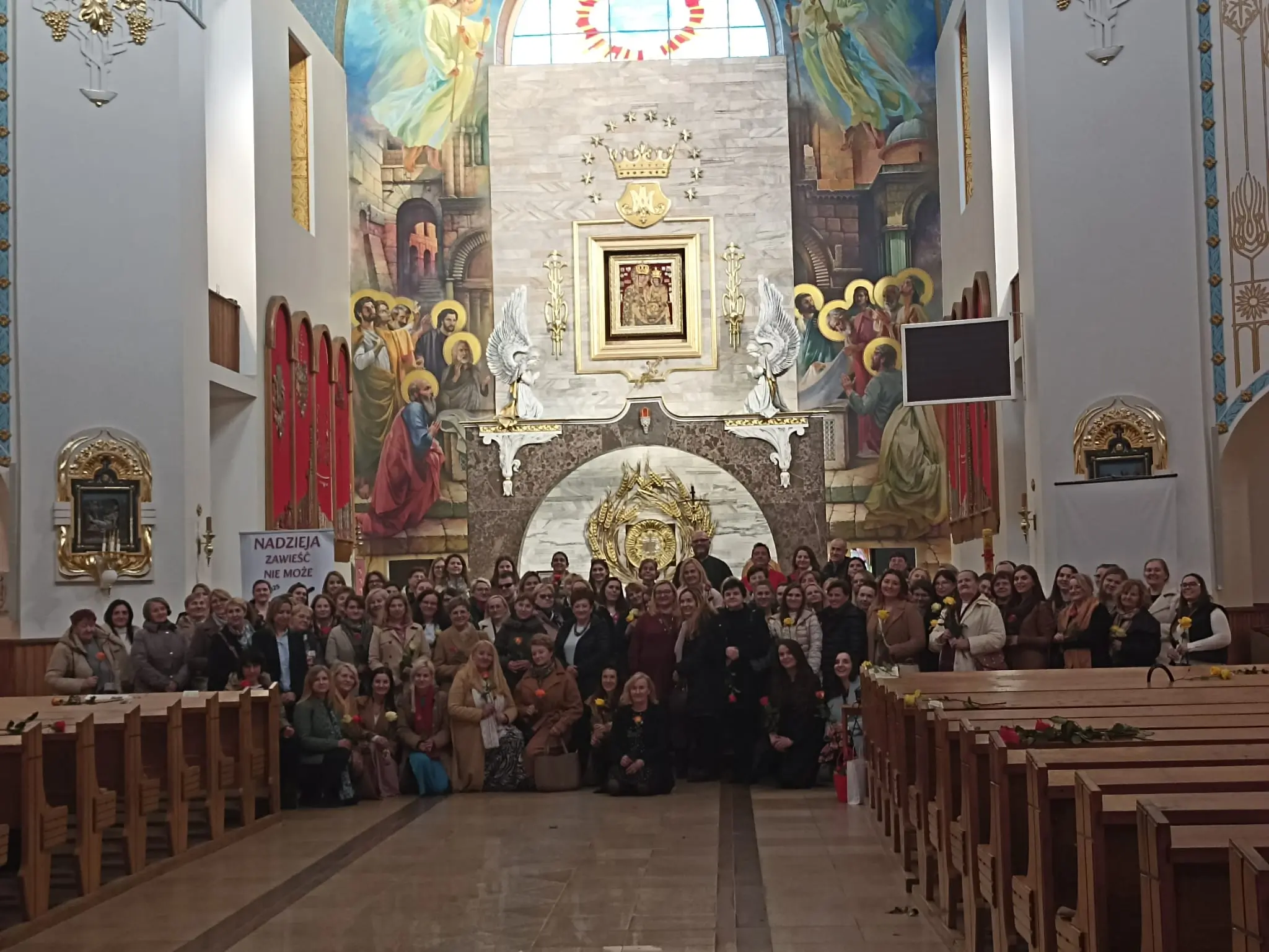Duża grupa osób zgromadzona w kościele podczas wydarzenia. W tle znajduje się kolorowa freska przedstawiająca scenę religijną. Na zdjęciu widoczna jest również tablica z napisem "NADZIEJA ZAWSZE NIE MOŻE".