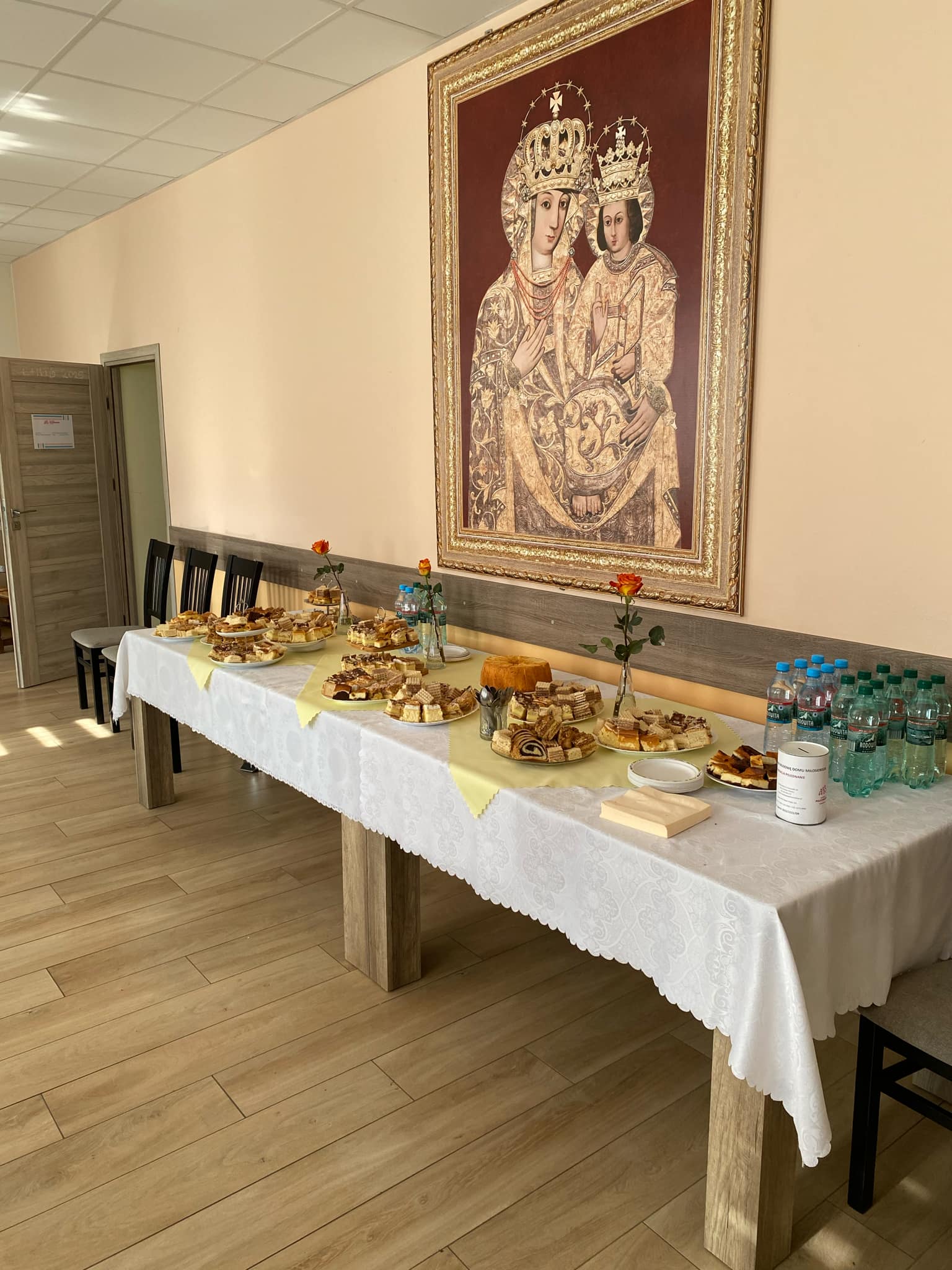 Na stole ustawione są podajniki z jedzeniem i napojami, a w tle znajduje się obraz przedstawiający dwóch świętych. Na stole wyłożona jest biała, wzorzysta tkanina.
