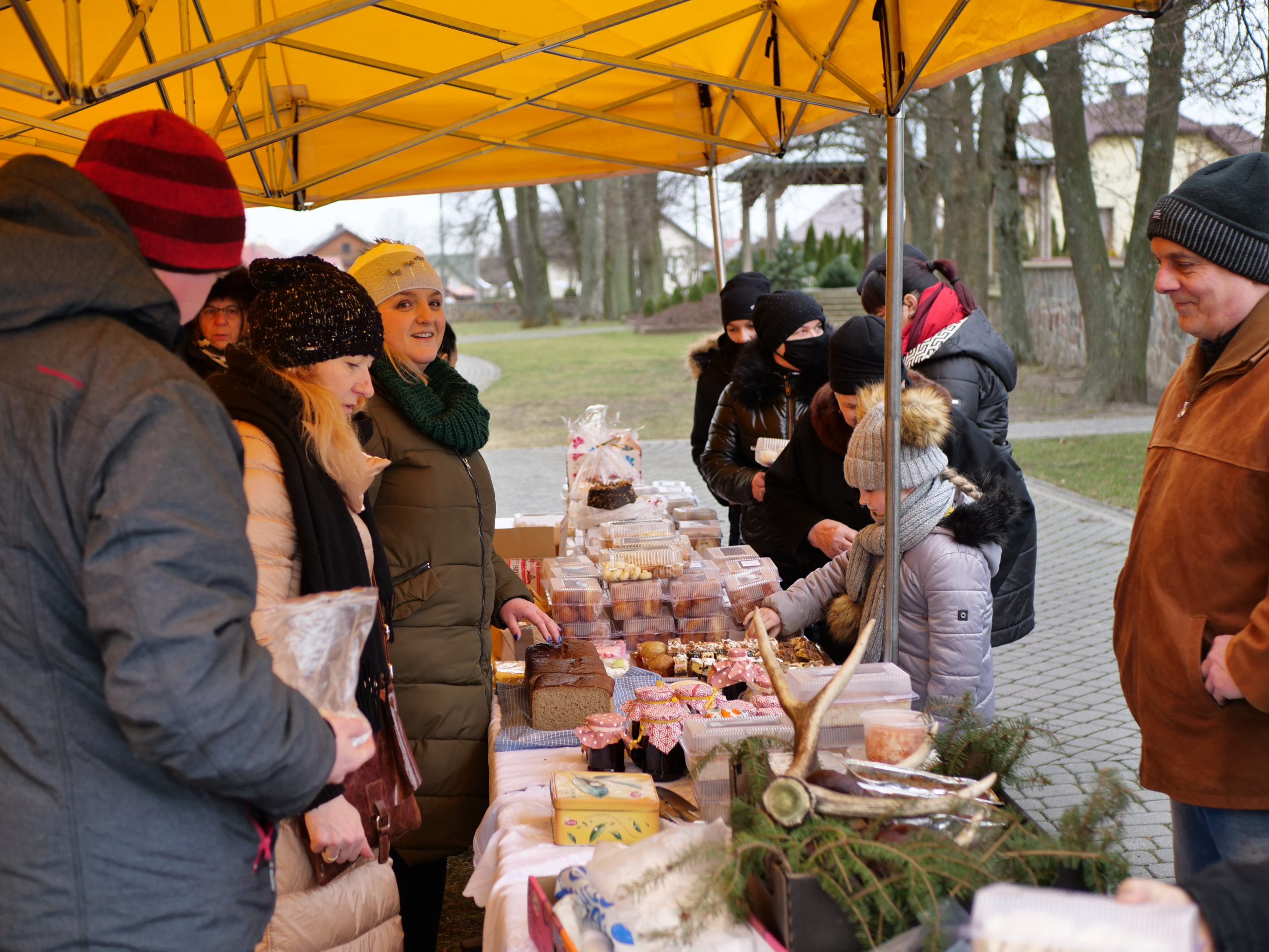 Na kiermaszu prezentowane są produkty spożywcze, takie jak ciastka i wędliny. Wokół stoiska zgromadzeni ludzie. Obywatel ubrany w czarny płaszcz stoi blisko stoiska.