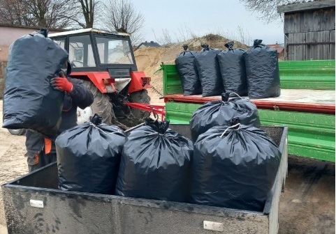 Zdjęcie przedstawia wózek transportowy wypełniony czarnymi workami plastycznymi, a osobę z rękawicami i pomarańczowymi rękawami trzymającą jeden z nich. Z tyłu widoczny jest traktor.