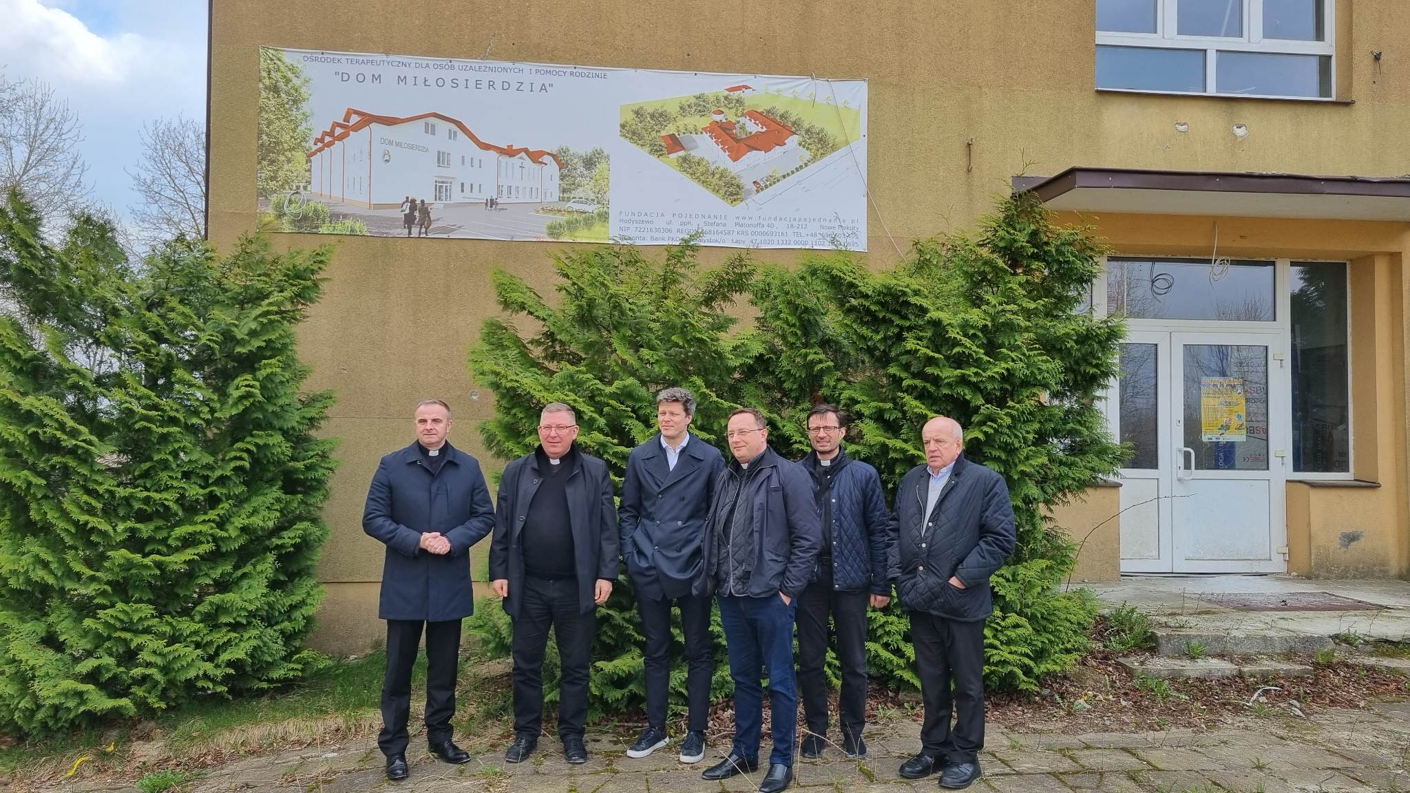 Sześciu mężczyzn stoi w grupce, ubranych w marynarki i spodnie. Na tle znajduje się plakat z projektem domu "Dom Miłosierdzia" oraz logo fundacji Pojednanie.