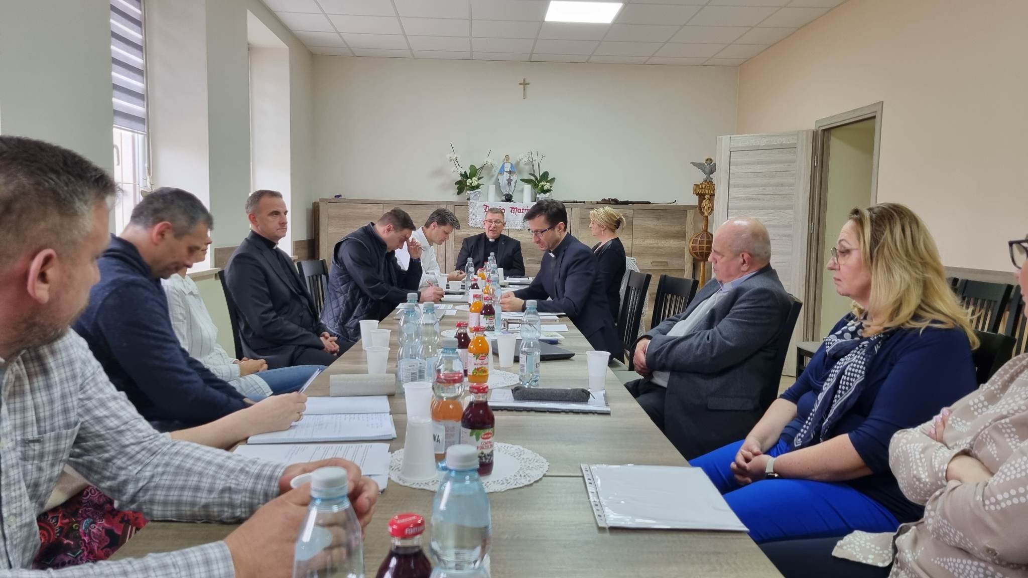 Grupa osób uczestniczy w spotkaniu w pokoju biurowym. Na stole znajdują się napoje i dokumenty.