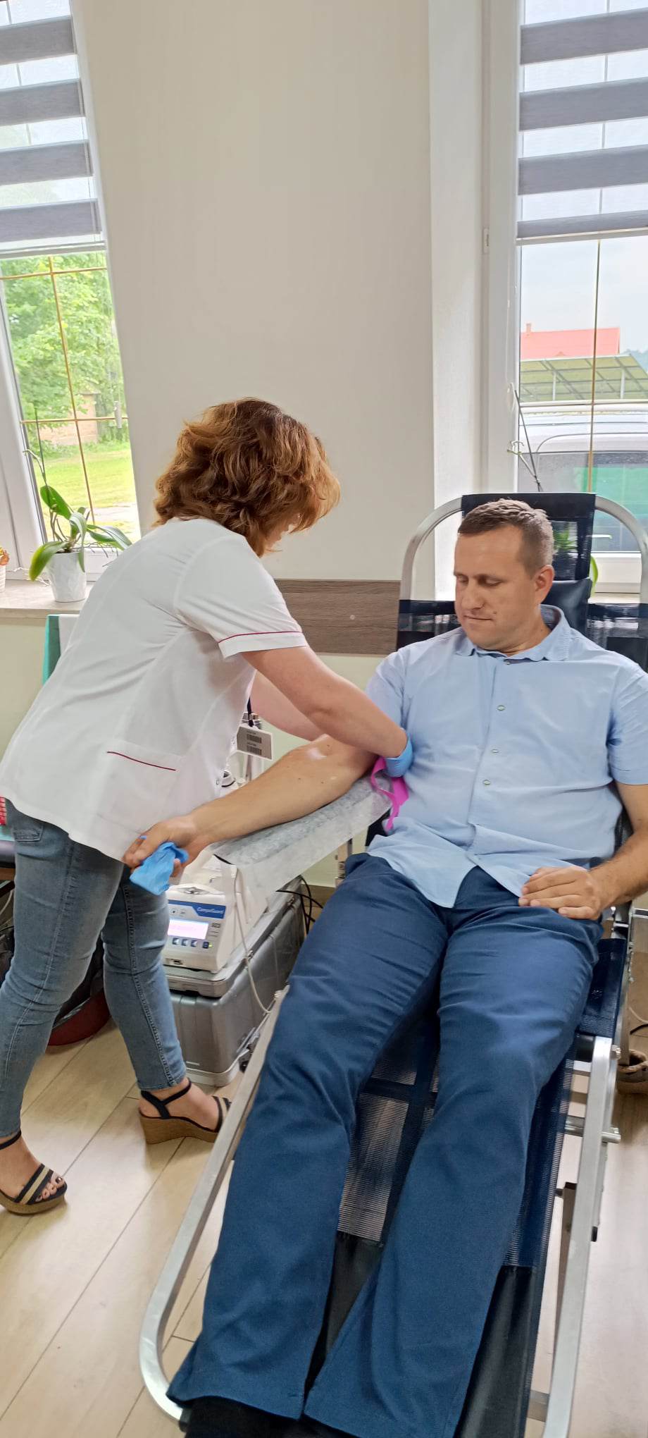 Zdjęcie przedstawia mężczyznę siedzącego w fotelu medycznym, podczas gdy pielęgniarka przygotowuje sprzęt do pobrania krwi. W tle widoczna szafka i okno z widokiem na zewnątrz.