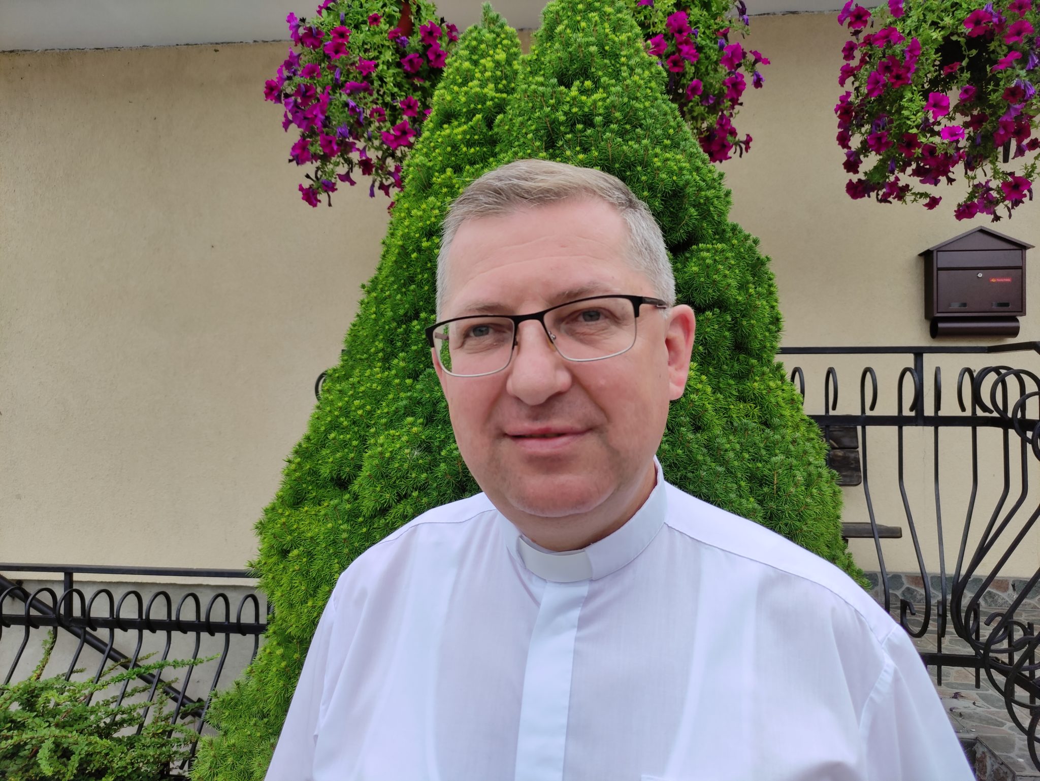 Fotografia osoby w białych szatach kapłańskich z okularami. W tle zielone krzewy i kwiaty, a także skrzynka pocztowa.