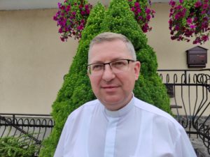 Mężczyzna w białym szatach kościelnych stoi na tle zielonego krzewu i budynku z kwiatami. Zdjęcie służy do przedstawienia osoby, która jest związana z działalnością Stowarzyszenia Apostolstwa Katolickiego.
