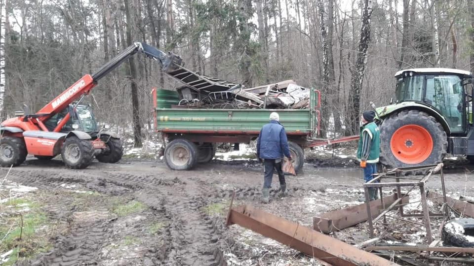 Na terenie robót widać przyczepę widlastą z przycumowanym do niej metalowym gruzem oraz ciągnik siodłowy. W tle znajduje się gęste drzewostan.